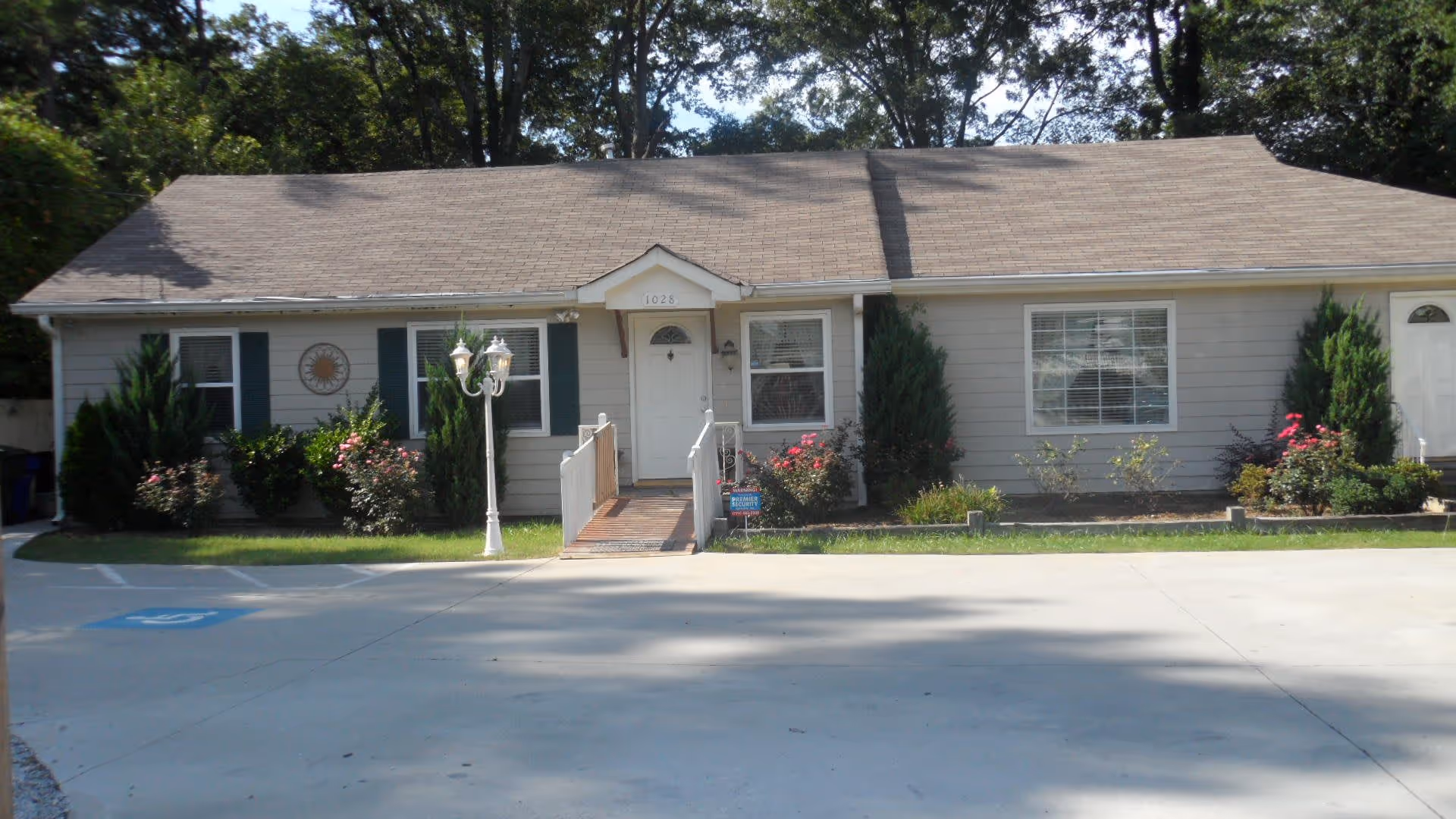 Single-story residential building with a beige exterior, white front door, and multiple windows. There is a small ramp leading to the entrance, surrounded by bushes and flowers. A white lamp post is positioned near the ramp. The building is set against a backdrop of tall trees and a clear sky. A parking area with a handicap parking space is visible in front.