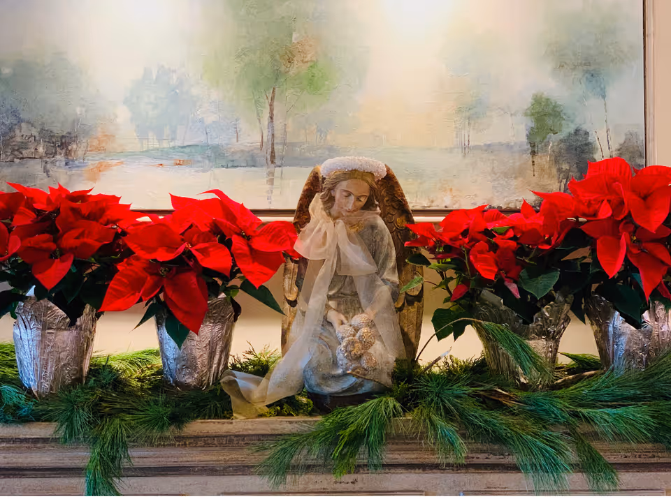 A holiday arrangement of red poinsettias and evergreen boughs flanking a seated angel figurine in front of a soft landscape painting.