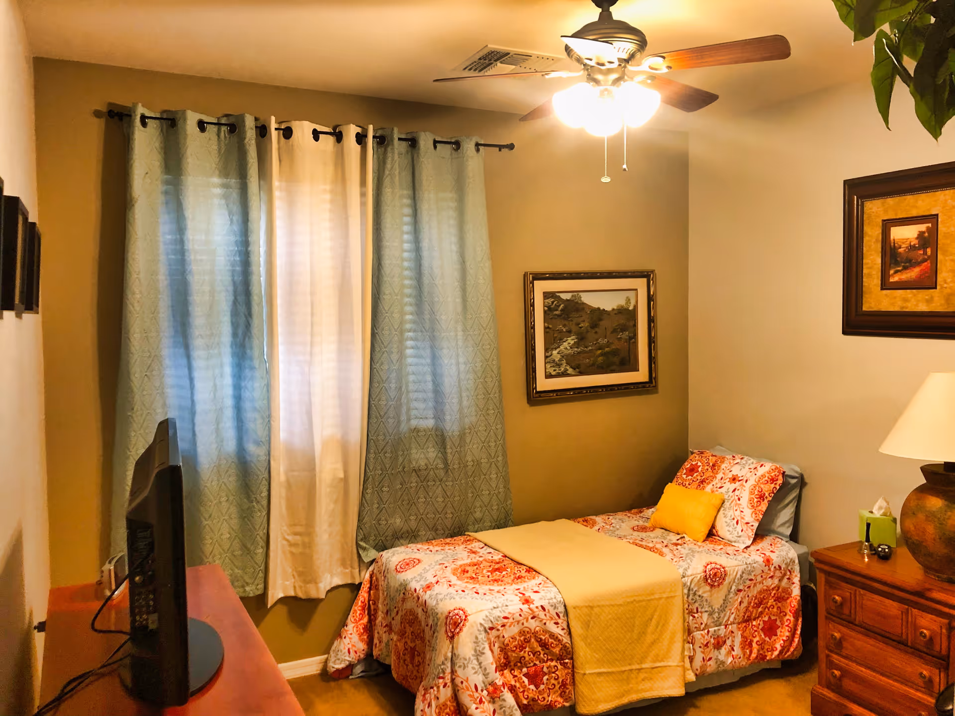 A cozy bedroom with a single bed covered in a colorful patterned bedspread and a yellow throw blanket. The room has beige walls, a ceiling fan with lights, a wooden dresser with a lamp and tissue box, and two framed paintings on the walls. A TV is placed on a wooden stand opposite the bed, and the window is covered with light blue and white curtains.