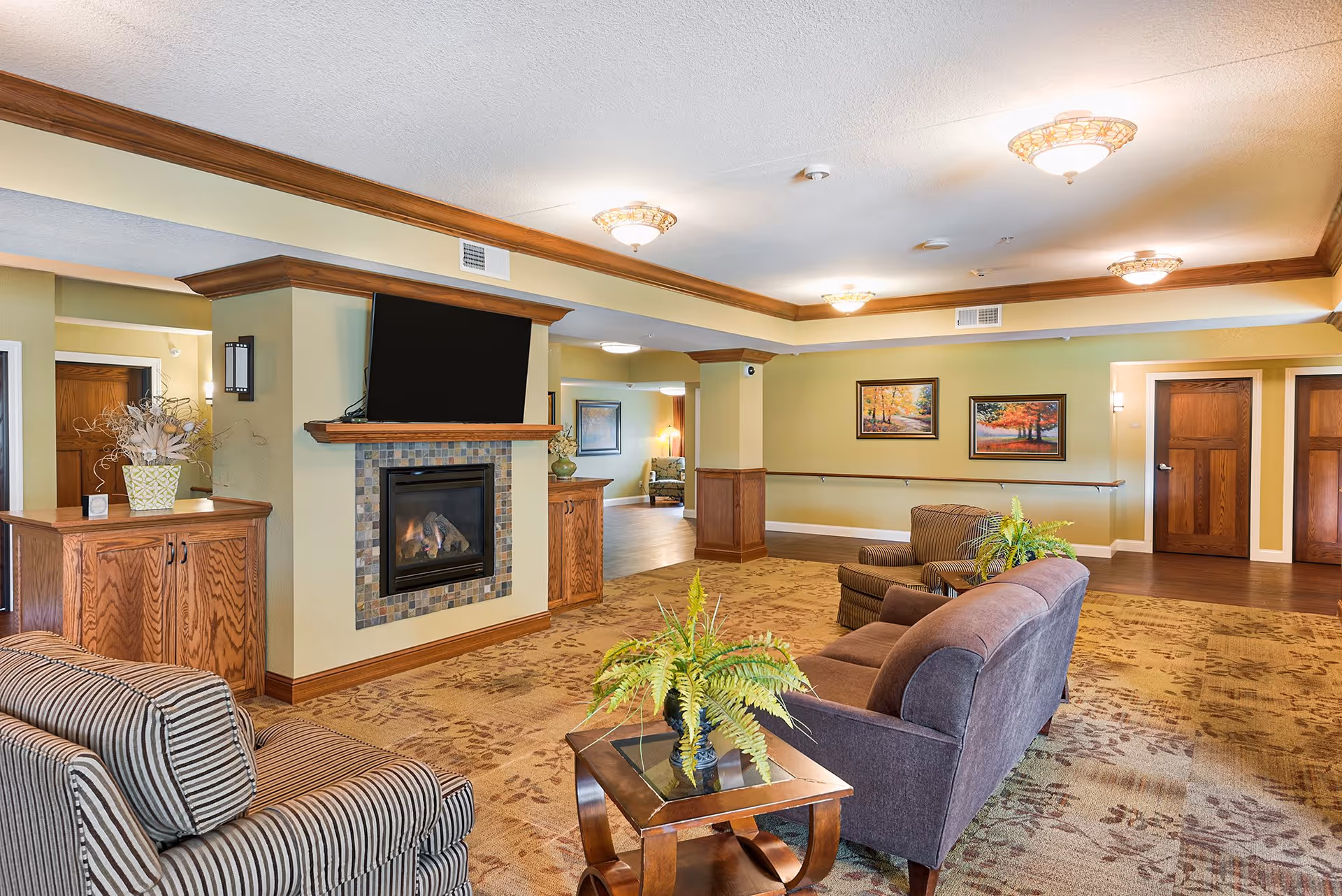 A cozy living room area in a senior living facility featuring a fireplace with a mounted TV above it, comfortable armchairs and a sofa arranged around a small wooden coffee table with a potted fern. The room has warm lighting, framed artwork on the walls, and wooden doors leading to other rooms.