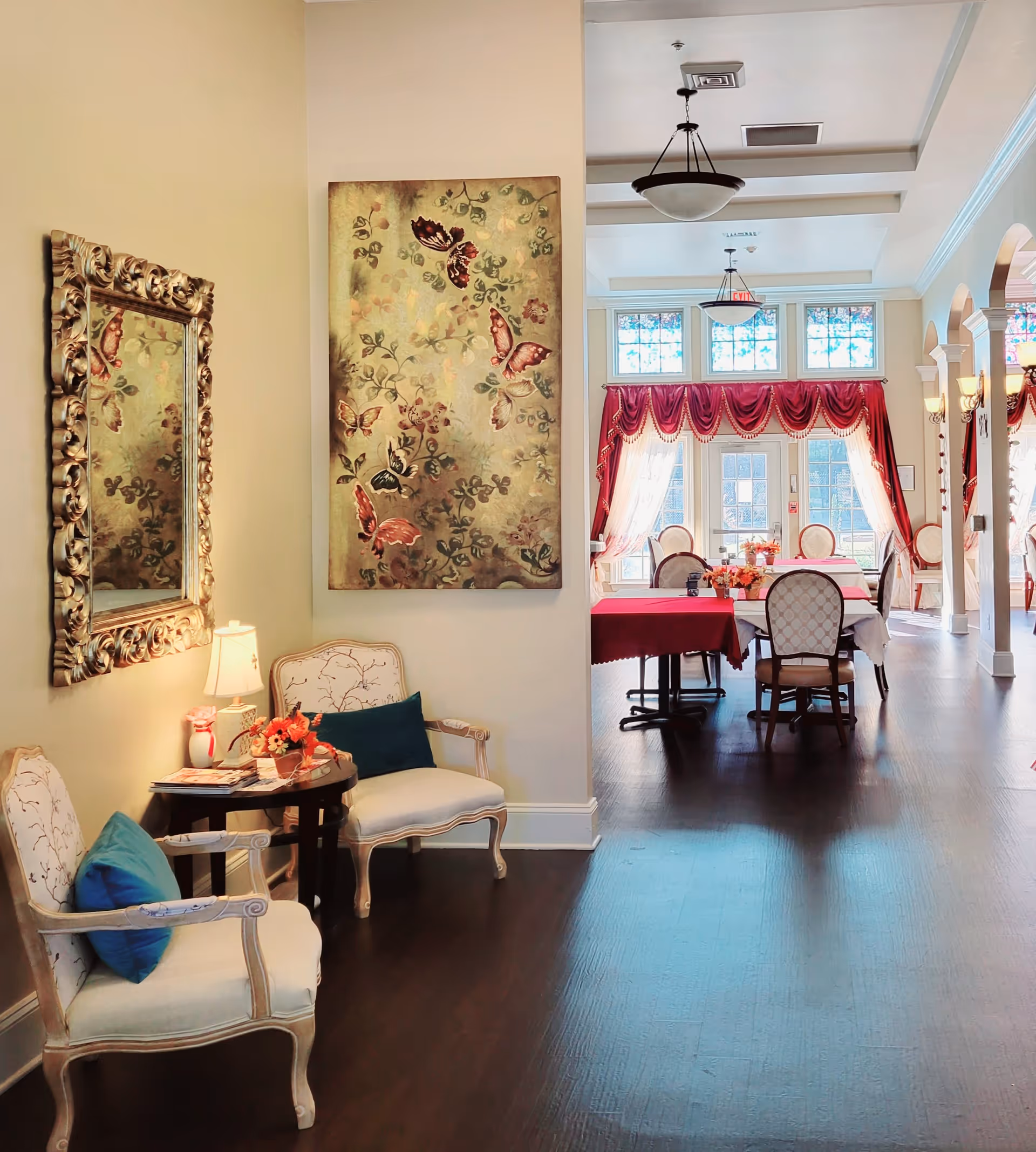 A bright and elegant interior space featuring a small seating area with two upholstered chairs with blue cushions, a round side table with a lamp and flower arrangement, a large ornate mirror, and a butterfly-themed wall art. In the background, a dining area with tables covered in red and white tablecloths, chairs, large windows with red drapes, and ceiling light fixtures is visible.