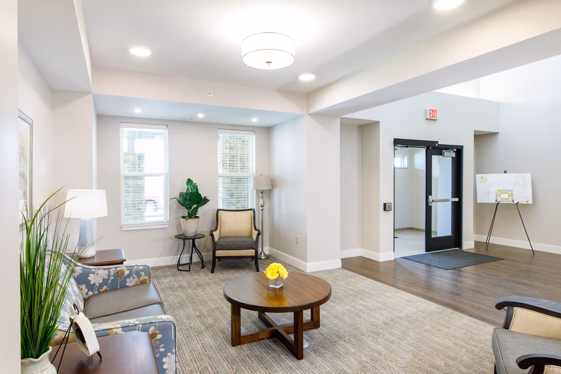 A bright and spacious assisted living facility common area with a floral patterned sofa, two armchairs, a round wooden coffee table with a small vase of yellow flowers, a floor lamp, and two windows letting in natural light. The room has light-colored walls, a carpeted floor, and a doorway leading to another area with a glass door and an exit sign above it. A plant and a framed picture add decorative touches.