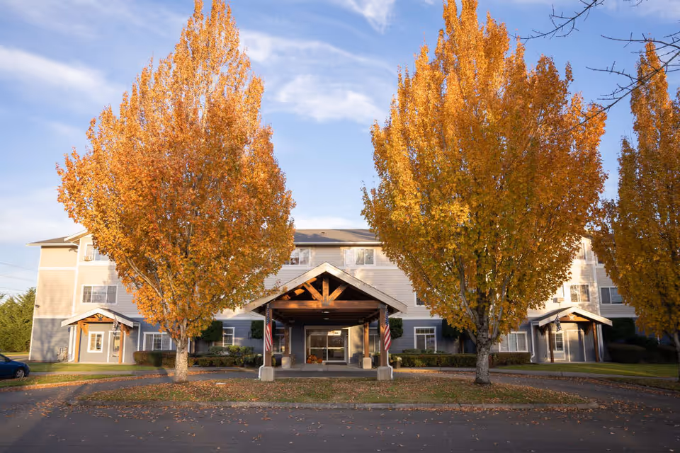 Front exterior view of St Michael's Assisted Living facility with a covered entrance, two large trees with orange autumn leaves, and a clear blue sky.