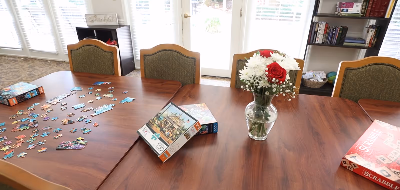 A wooden table with a partially completed jigsaw puzzle, puzzle boxes, a Scrabble board game, and a vase of red and white flowers. The table is surrounded by chairs with green patterned upholstery. In the background, there are large windows with blinds and a bookshelf with books and baskets.