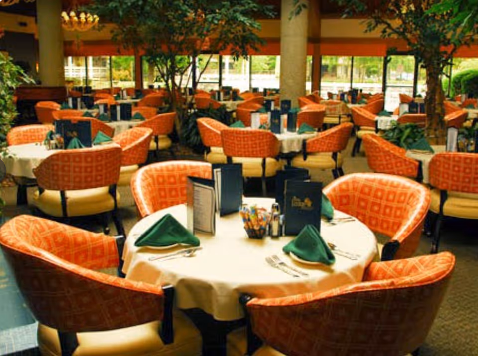 A spacious dining room with multiple round tables covered with white tablecloths, each set with green folded napkins, menus, and silverware. The chairs are upholstered in orange patterned fabric with cream-colored seats. Large windows and indoor plants provide a bright and inviting atmosphere.