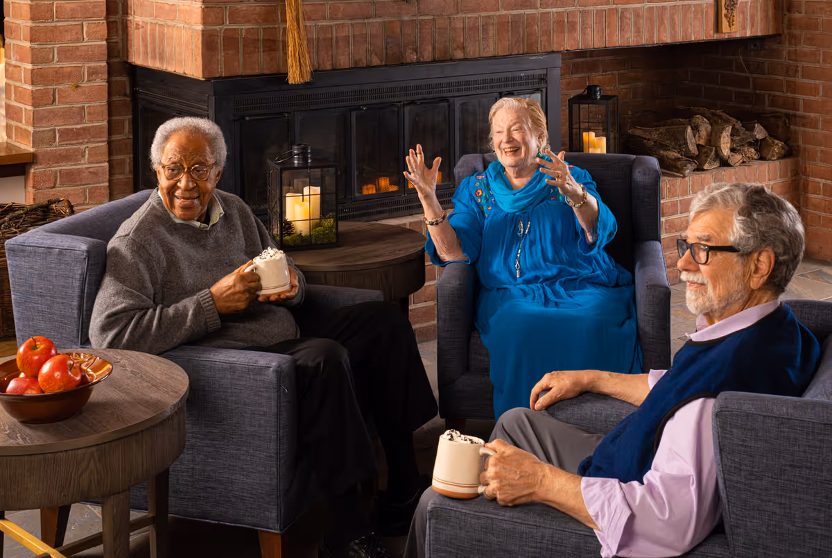 Three elderly people sitting in armchairs around a fireplace in a cozy room. Two men and one woman are enjoying hot beverages topped with whipped cream. The woman in a blue dress is animatedly talking, while the men listen and smile. A bowl of apples is on a nearby wooden table.