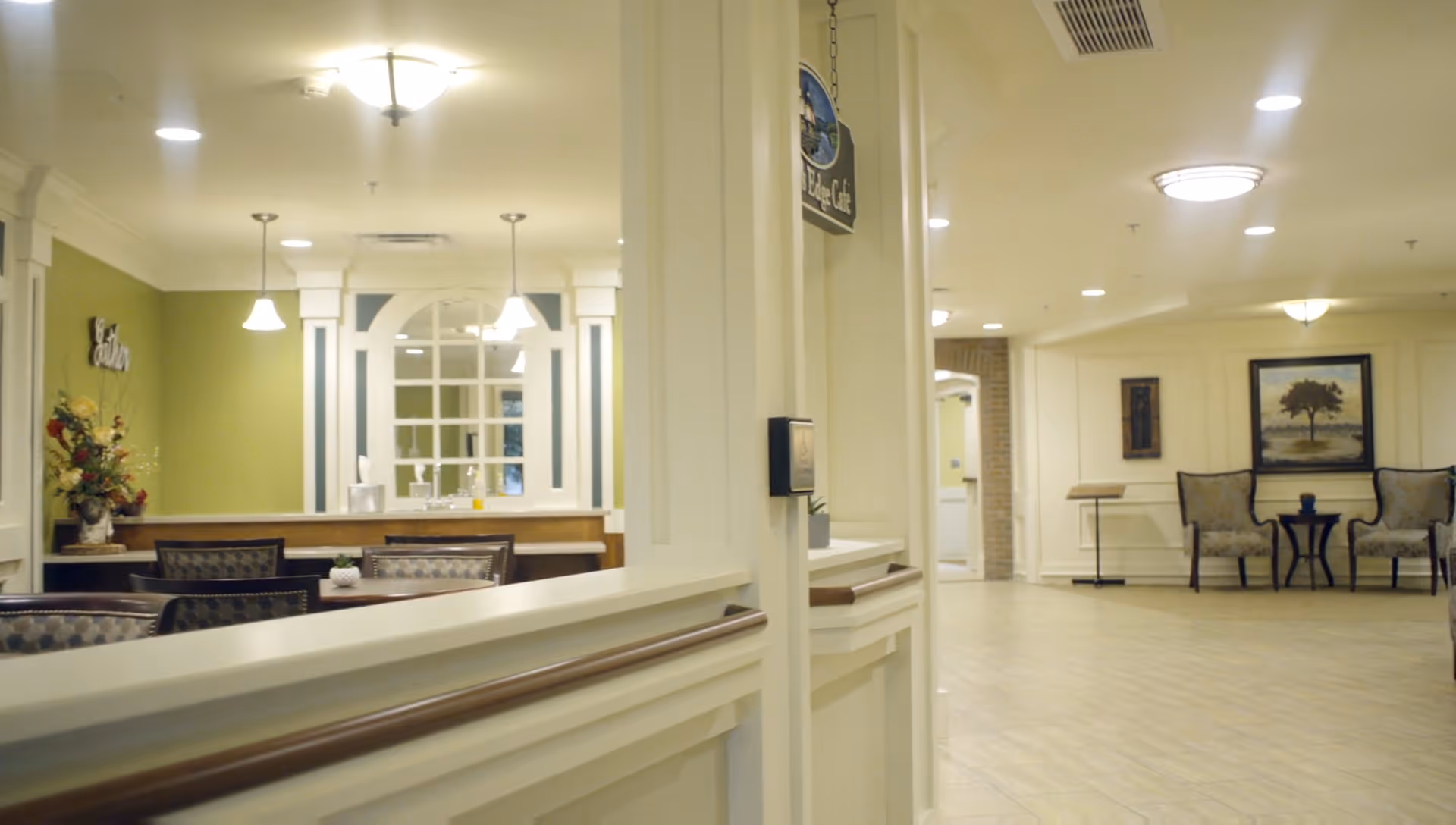 Interior view of a senior living facility showing a dining area with tables and chairs on the left, and a sitting area with two armchairs and a small table with a painting on the wall in the background. The walls are light-colored with decorative molding, and the floor is tiled. Ceiling lights illuminate the space.