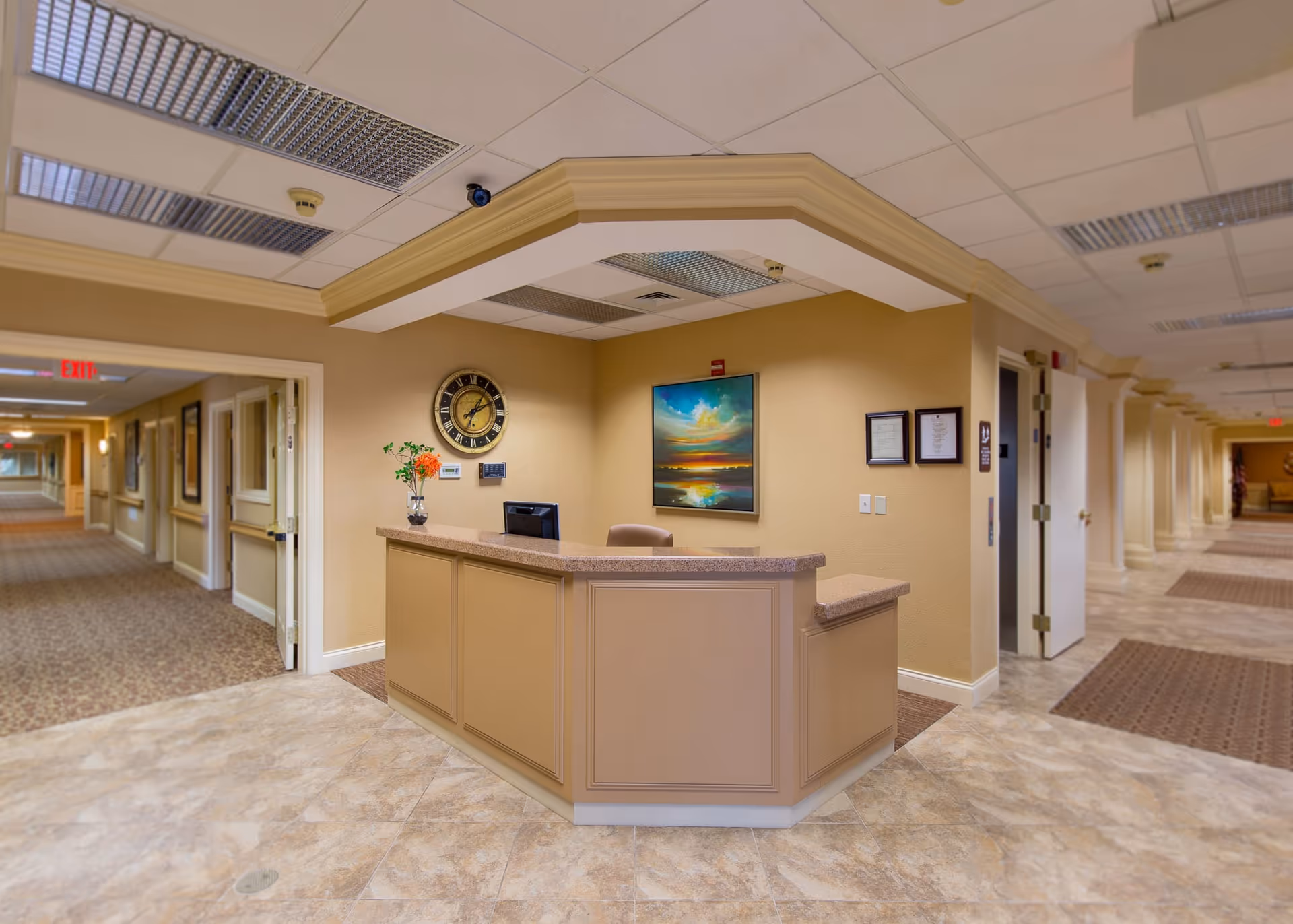 Reception desk in a senior living facility lobby with a wall clock, artwork, and corridors extending down both sides.