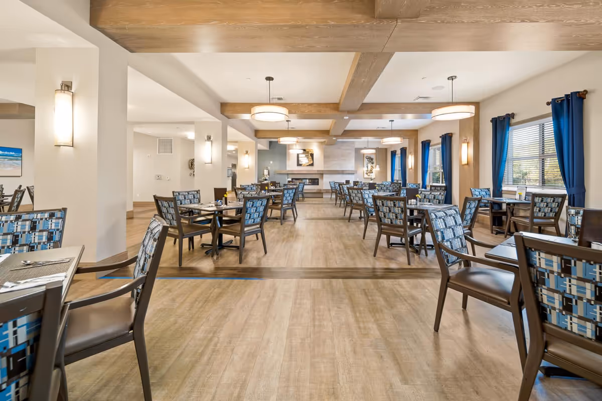 A spacious dining room with multiple tables and chairs arranged neatly. The chairs have patterned blue upholstery, and the room features wooden beams on the ceiling, modern pendant lights, and large windows with blue curtains allowing natural light to enter. The floor is wooden, and the walls are painted in light colors with some decorative elements.