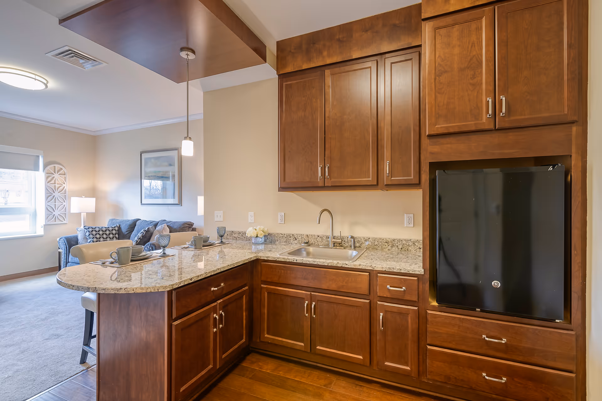 Open kitchen with dark wood cabinets, a granite countertop island with place settings and a sink, opening onto a living area with a couch and lamp.