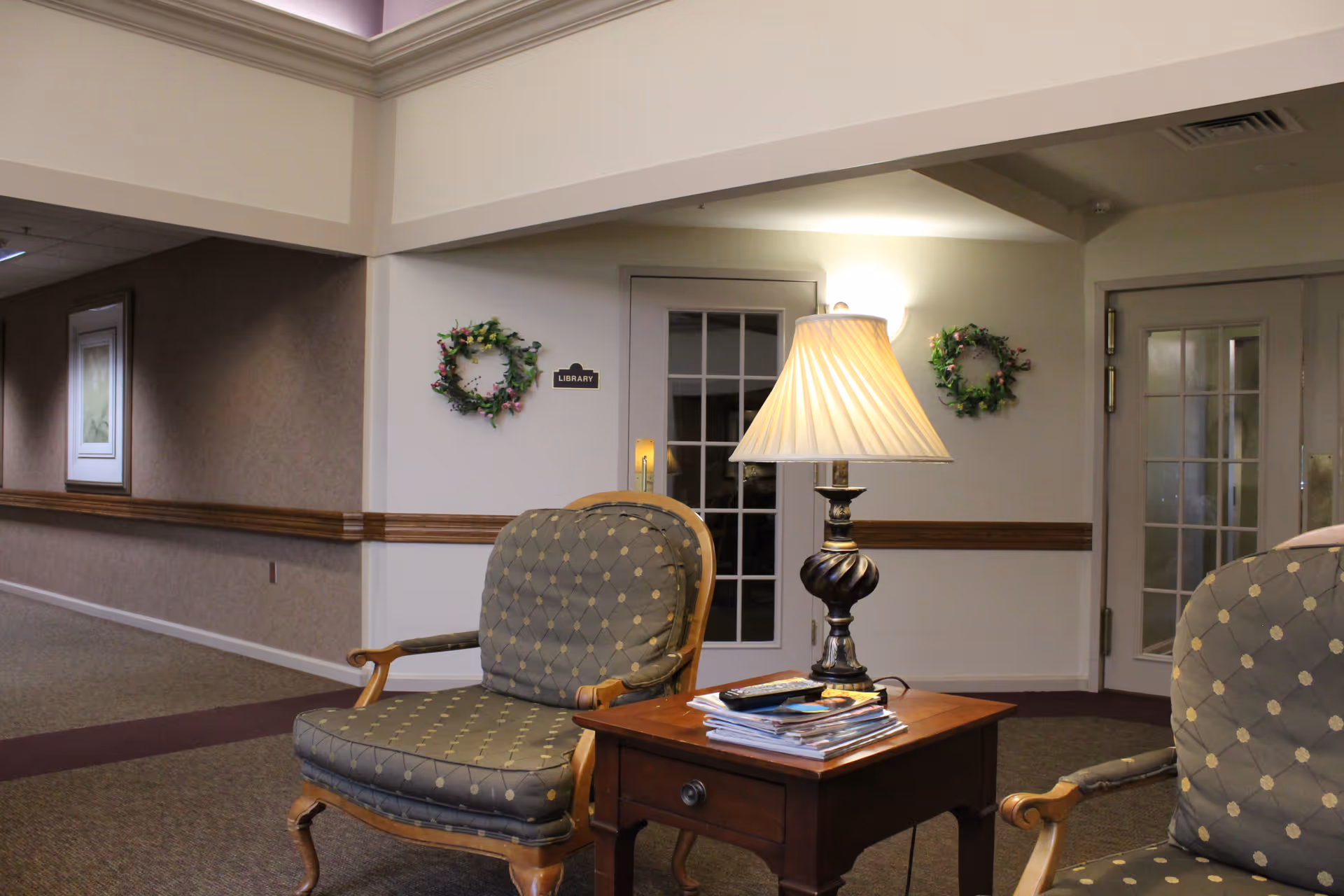 A cozy seating area in an assisted living facility hallway with two upholstered armchairs featuring a diamond pattern, a wooden side table with a lamp and magazines, and a door labeled 'Library' decorated with floral wreaths on either side.