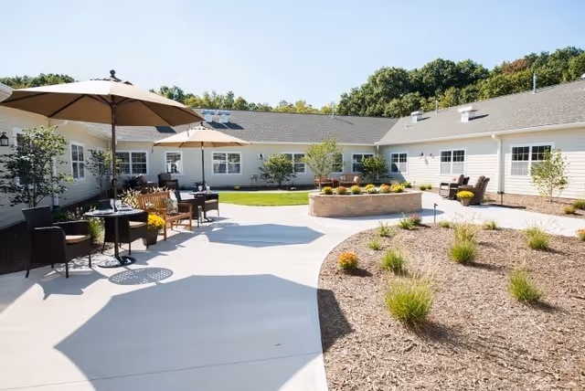 Outdoor courtyard area at The Residence at Glastonbury featuring a paved walkway, patio tables with umbrellas, chairs, a raised circular planter with flowers, and surrounding single-story building with windows under a clear blue sky.