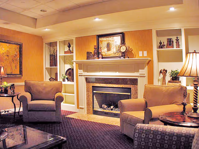 A cozy living room area with two beige armchairs, a fireplace with a mantle decorated with a clock and framed artwork, built-in shelves with decorative items, a side table with a lamp, and warm lighting.