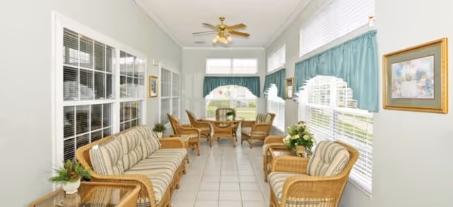 A bright sunroom with large windows on three sides, furnished with wicker chairs and sofas with striped cushions, a small round table with chairs at the far end, potted plants, and teal curtains on the windows.