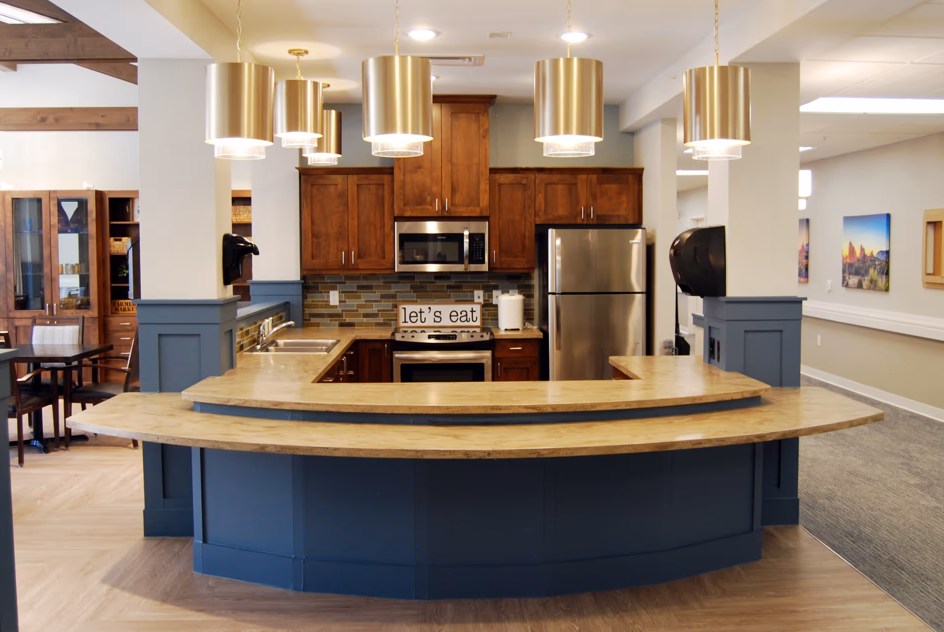A modern kitchen area in a senior living facility featuring a curved wooden countertop with blue paneling, stainless steel refrigerator and microwave, wooden cabinets, a stove with a sign that reads 'let's eat', and four gold pendant lights hanging from the ceiling. Adjacent dining area with tables and chairs is partially visible.