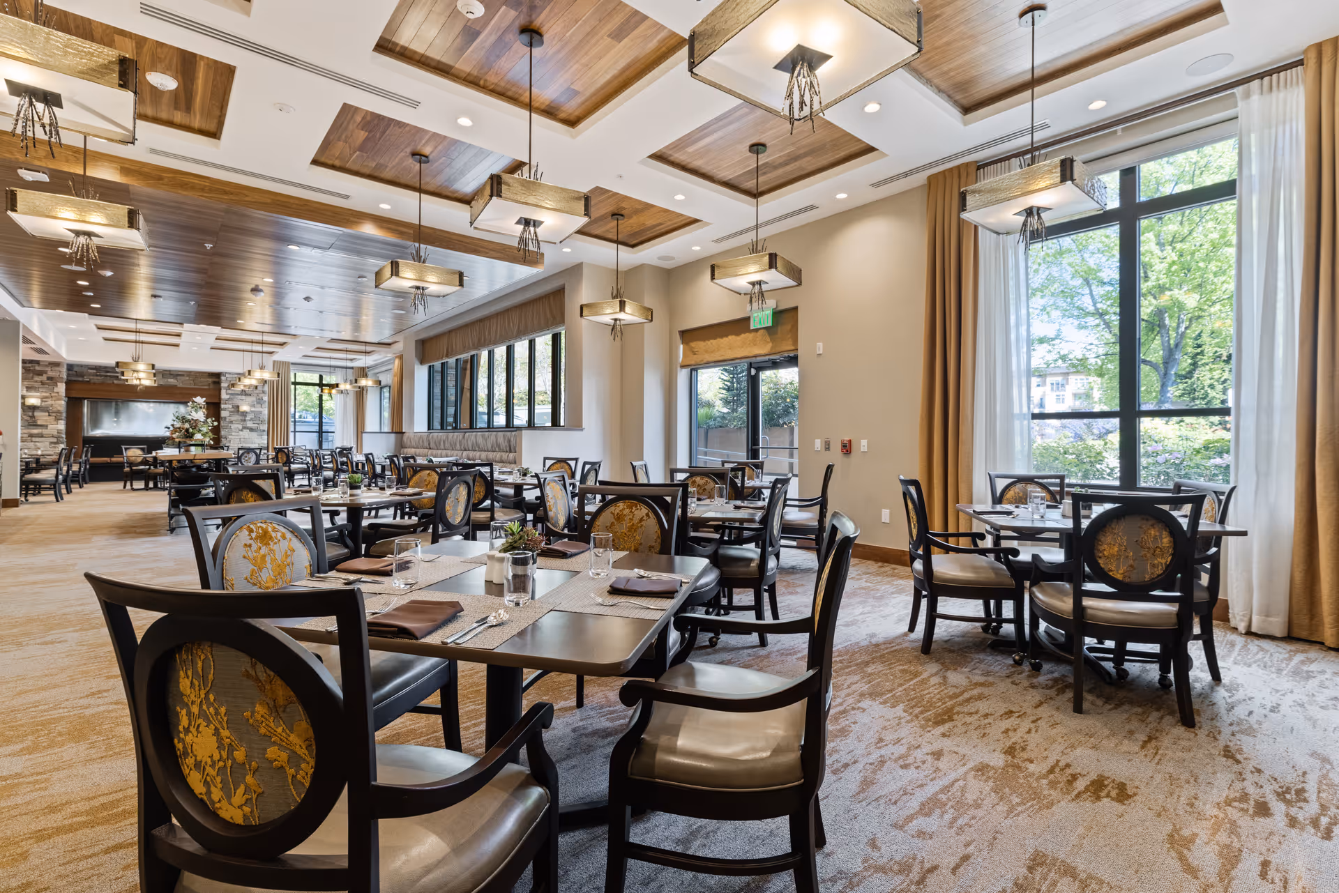 A spacious, well-lit dining room with multiple tables and chairs arranged neatly. The room features large windows with beige curtains allowing natural light to fill the space. The ceiling has wooden panels with modern rectangular light fixtures hanging down. The tables are set with placemats, napkins, glassware, and small decorative plants.