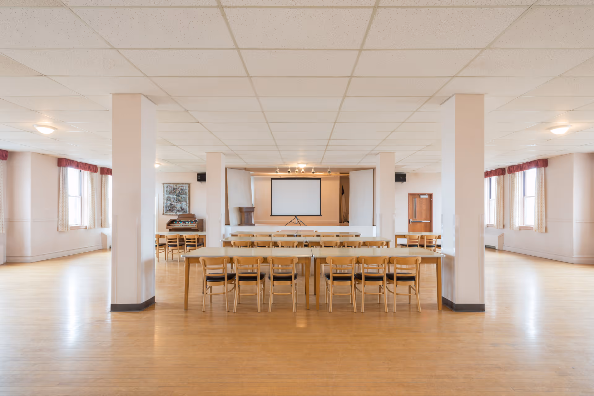 Spacious multipurpose meeting room with rows of tables and chairs facing a projection screen, wooden floors, pillars, and windows with curtains.