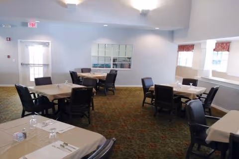 A dining room with several tables covered with beige tablecloths, each set with placemats, napkins, utensils, and glasses. The room has carpeted flooring, white walls, and windows with red patterned valances letting in natural light. There is a door and a window on the far wall.