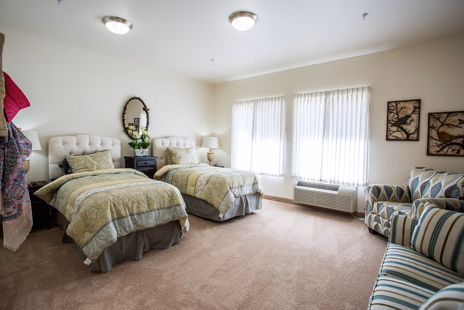 A bright and clean bedroom with two twin beds featuring patterned bedspreads, a nightstand with a lamp and a vase of flowers between the beds, two windows with white curtains, and a seating area with a striped couch and a patterned armchair. The walls are decorated with two bird-themed artworks and an oval mirror above the nightstand.