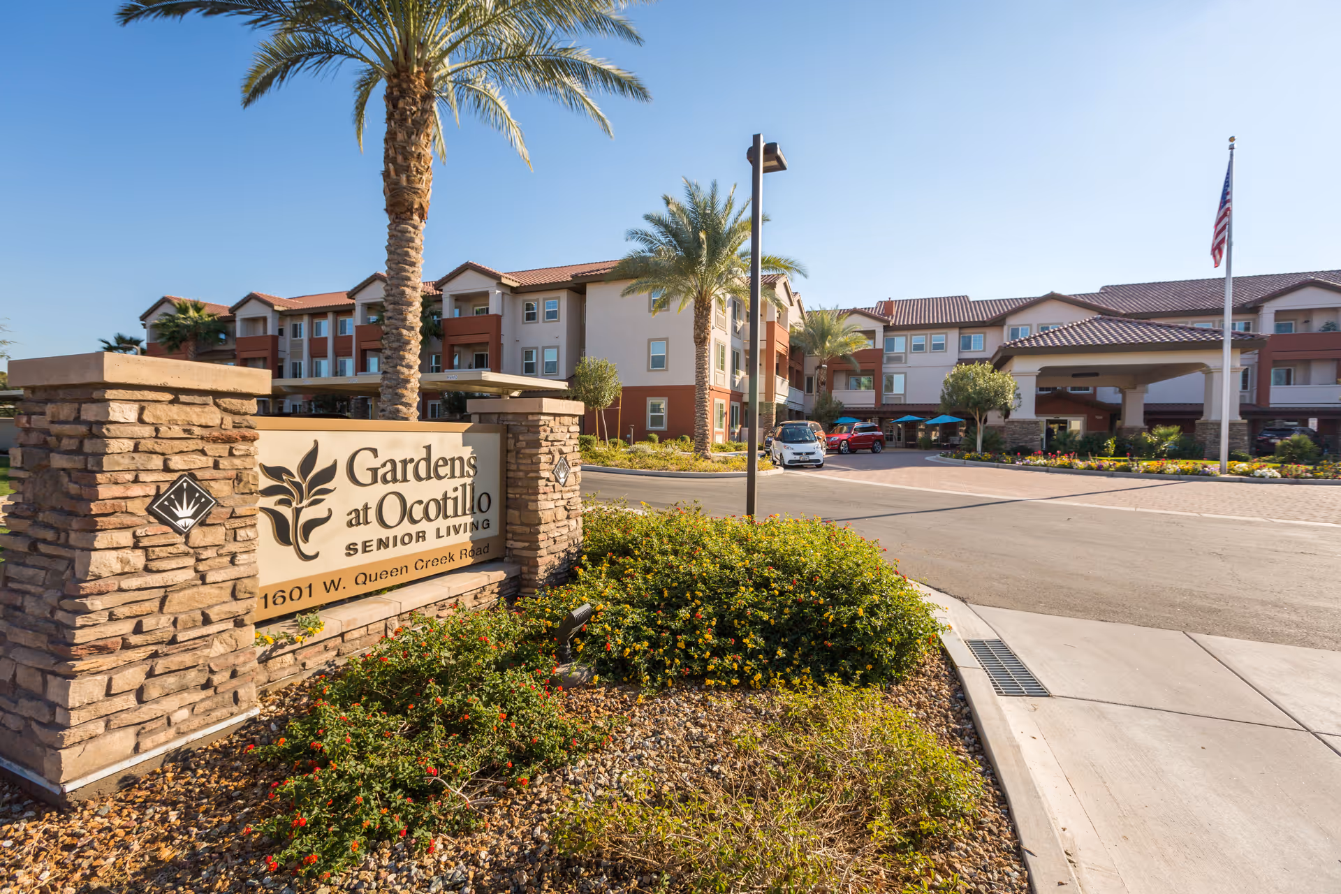 Exterior view of the Gardens at Ocotillo senior living facility showing the main entrance sign, landscaped garden with bushes and palm trees, a driveway with parked cars, and a multi-story building with balconies under a clear blue sky.