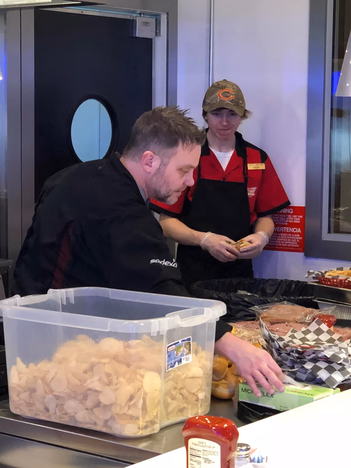 Two food service workers preparing sandwiches behind a counter with a large plastic tub of chips and condiments.