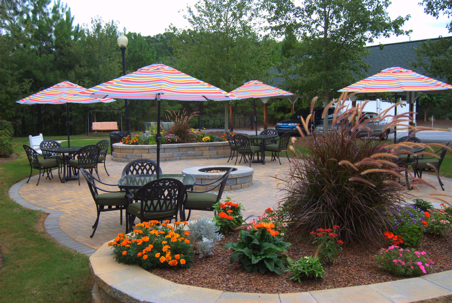 Outdoor patio area with several round tables and chairs, each shaded by colorful striped umbrellas. The patio is surrounded by landscaped flower beds with various plants and flowers, and there is a fire pit in the center. Trees and greenery are visible in the background.