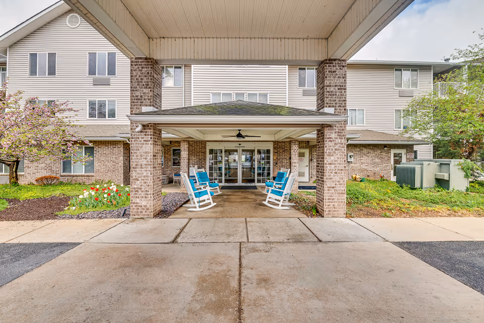 Entrance of a senior living facility with a covered driveway supported by brick pillars. Four blue cushioned rocking chairs are placed under the covered area. The building has beige siding and multiple windows. There are flower beds with tulips and a blossoming tree on the left side.