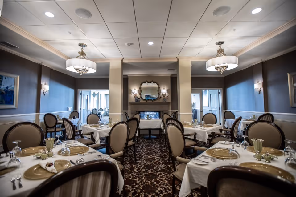 Elegant dining room with multiple tables set with white tablecloths, gold plates, glassware, and silverware. The room features upholstered chairs, a patterned carpet, a fireplace with a decorative mirror above it, and soft lighting from ceiling fixtures and wall sconces.