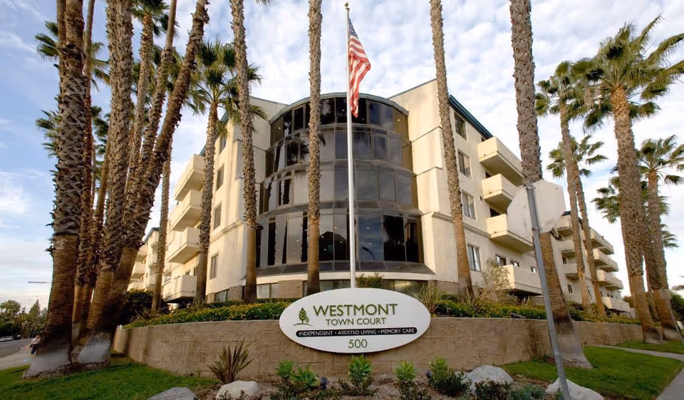 Exterior view of Westmont Town Court, a multi-story senior living facility surrounded by tall palm trees and landscaping, with an American flag flying in front and a sign indicating independent, assisted living, and memory care services.