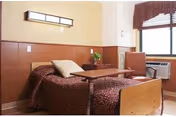 A single bed with a brown patterned bedspread and a beige pillow in a room with wood-paneled walls. Next to the bed is a small adjustable table, a brown upholstered chair, a window with blinds and a maroon valance, and a wall-mounted air conditioning unit.