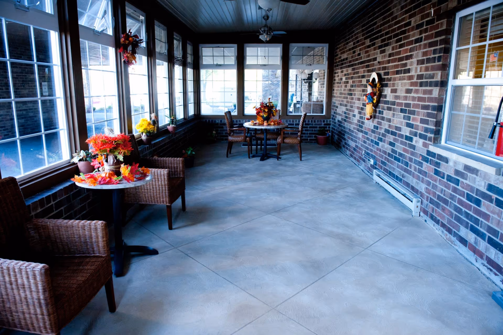 A bright enclosed porch area with large windows along one side and a brick wall on the other. The space features wicker chairs and small tables decorated with autumn-themed floral arrangements. The floor is tiled, and there is a decorative wreath hanging on the brick wall.