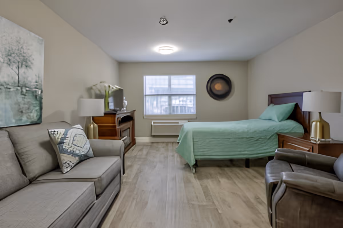 A senior living facility bedroom with a single bed covered in a green bedspread, a gray sofa with a decorative pillow, a brown armchair, a wooden nightstand with a lamp, a wooden dresser with a TV, and a window letting in natural light.