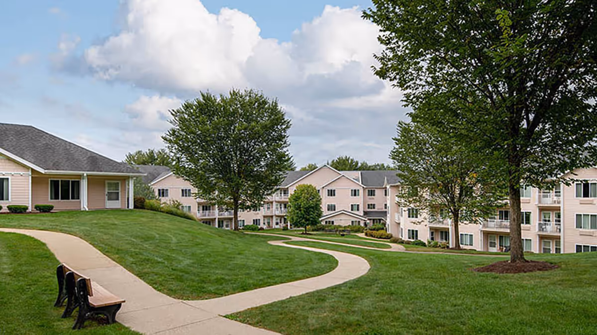 Outdoor view of Holiday Maple Suites featuring a winding concrete pathway through a well-maintained grassy area with several trees and benches. Surrounding the lawn are multi-story residential buildings with balconies under a partly cloudy sky.