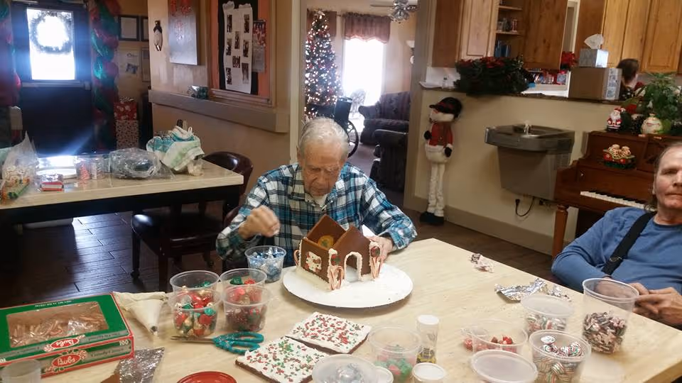 Two elderly men sitting at a table in a decorated room, one man is assembling and decorating a gingerbread house with various candies and icing, while the other man sits nearby. The room has holiday decorations including a Christmas tree, a snowman figure, and festive garlands.