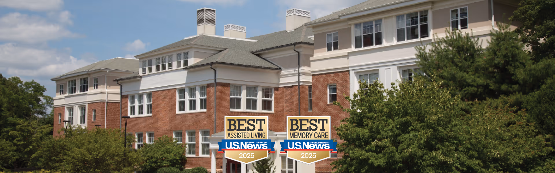 Brick and stucco senior living building facade with landscaping and two US News 'Best Assisted Living' and 'Best Memory Care' 2025 award signs in front.