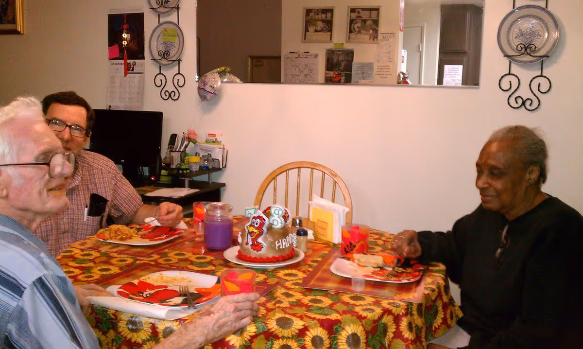 Three older adults sit around a sunflower-print dining table, eating and sharing a small birthday cake.