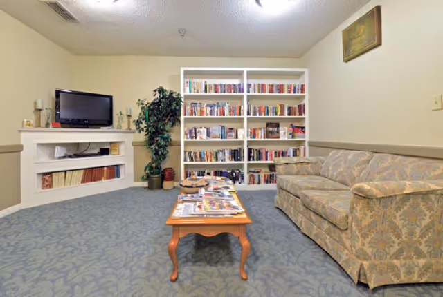 A cozy living room area with a patterned sofa on the right, a wooden coffee table with magazines on it in the center, a white bookshelf filled with books against the back wall, a potted plant next to the bookshelf, and a TV placed on a white built-in shelf on the left side of the room.