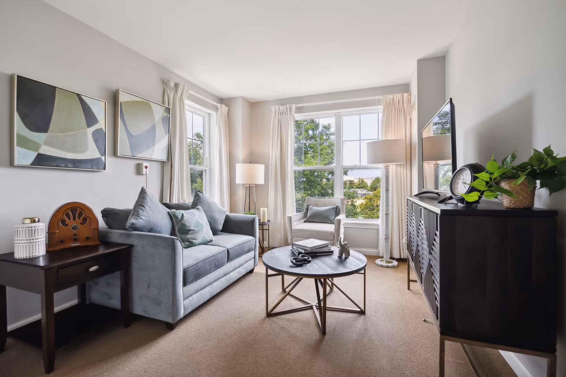 A bright living room with a large window letting in natural light. The room features a gray sofa with cushions, a round coffee table with books and decorative items, a single armchair near the window, a dark wooden side table with a vintage radio and candle, and a dark wooden cabinet with a clock and potted plant. Two abstract paintings hang on the wall above the sofa.