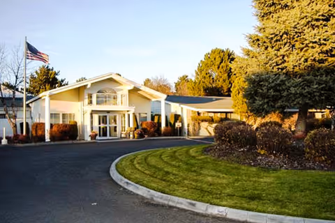 Front exterior view of a single-story building with a white facade, large windows, and a covered entrance. There is a curved driveway in front, well-maintained green lawn, bushes, and trees. An American flag is flying on a flagpole to the left of the building.