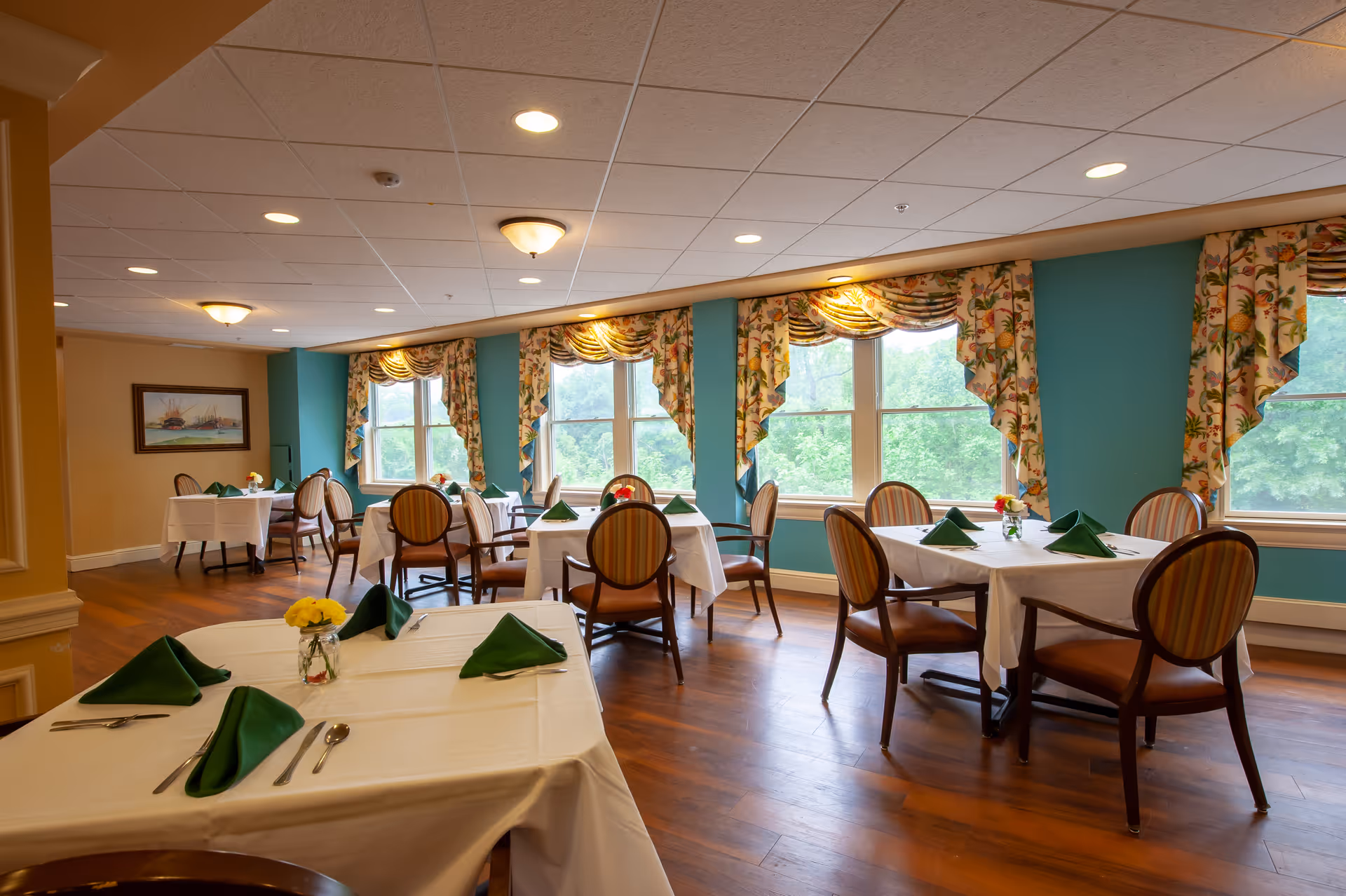 A bright dining room with several tables covered in white tablecloths, each set with green folded napkins, silverware, and small flower vases. The room has large windows with floral curtains, teal-colored walls, and wooden flooring. The ceiling has recessed lighting and decorative light fixtures.