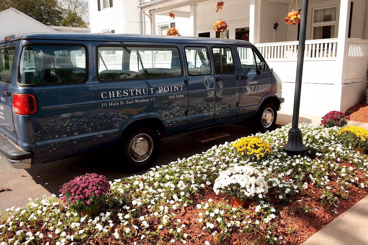 A blue van parked outside a white building with a porch decorated with hanging flower baskets. The van has the text 'CHESTNUT POINT 171 Main St. East Windsor, CT 292-5394' on its side. In front of the van is a flower bed with white, yellow, and purple flowers, and a black lamp post is visible near the flower bed.