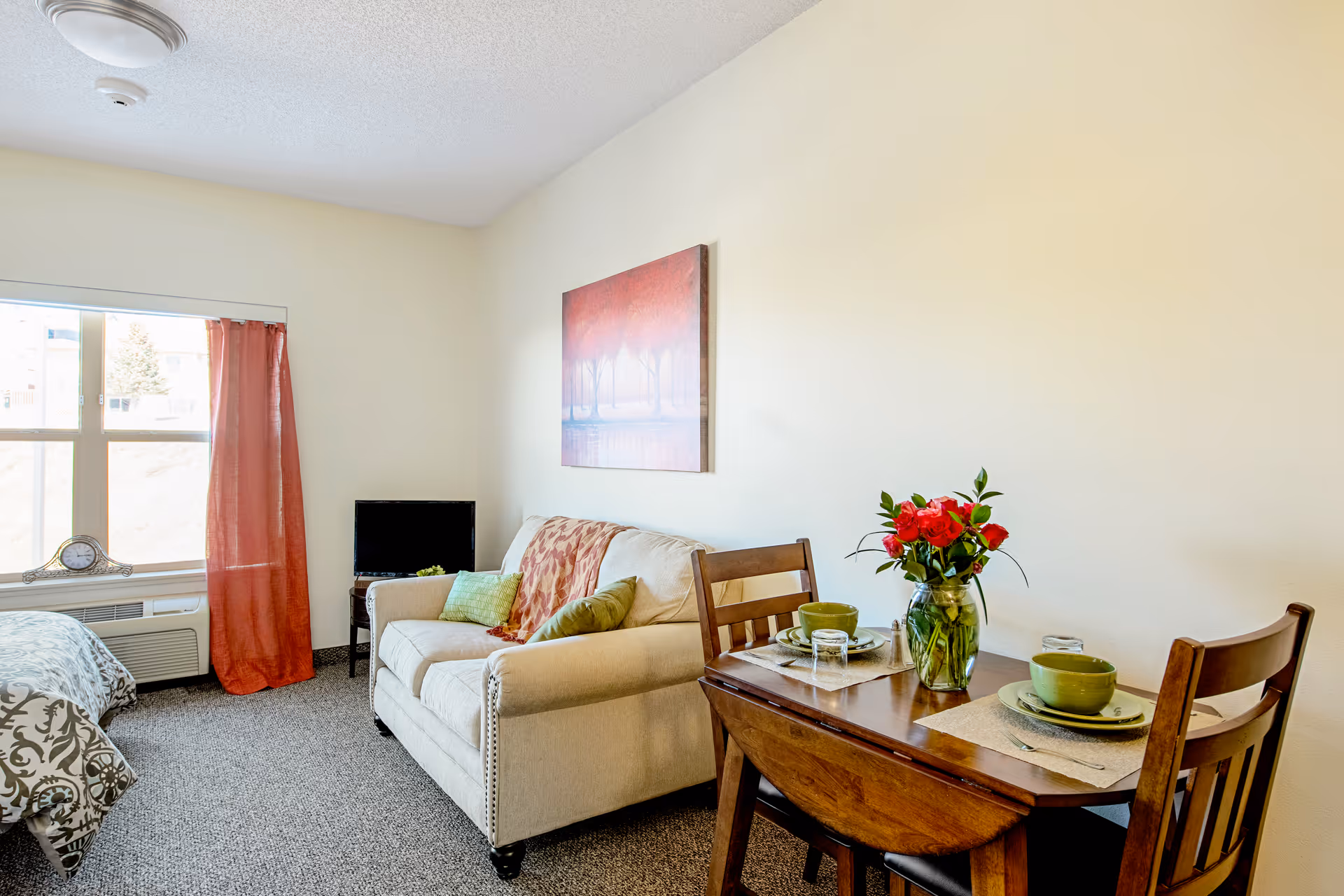 A cozy living space with a beige sofa adorned with green and patterned cushions, a small wooden dining table set for two with green dishes and a vase of red flowers, a small TV on a stand near a window with red curtains, and part of a bed with patterned bedding visible on the left side.