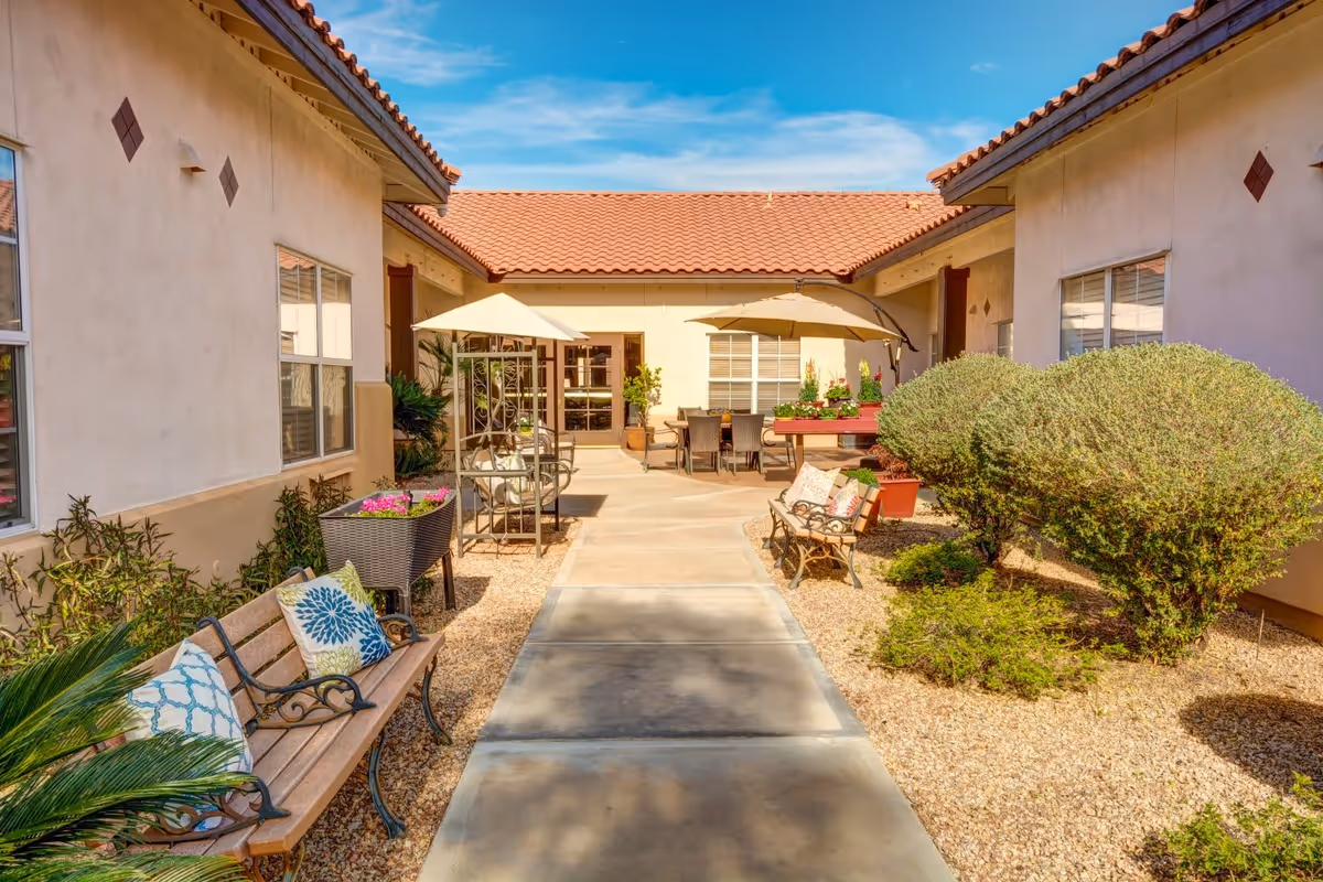 Outdoor courtyard area with a paved walkway flanked by benches with decorative pillows, potted plants, and trimmed bushes. The courtyard is surrounded by buildings with beige walls and red tile roofs under a clear blue sky. There are umbrellas providing shade over seating areas near the building entrance.