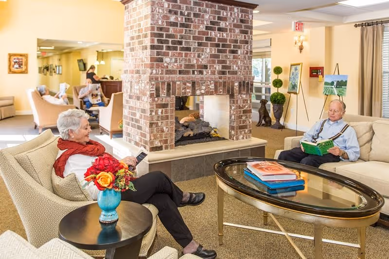 A cozy senior living facility common area with a brick fireplace in the center. Two elderly individuals are seated on comfortable chairs and sofas, reading books. A glass coffee table with books on it is in front of them, and a small table with a vase of colorful flowers is nearby. The room is warmly lit with beige walls and carpeted floors, and there are decorative plants and paintings in the background.