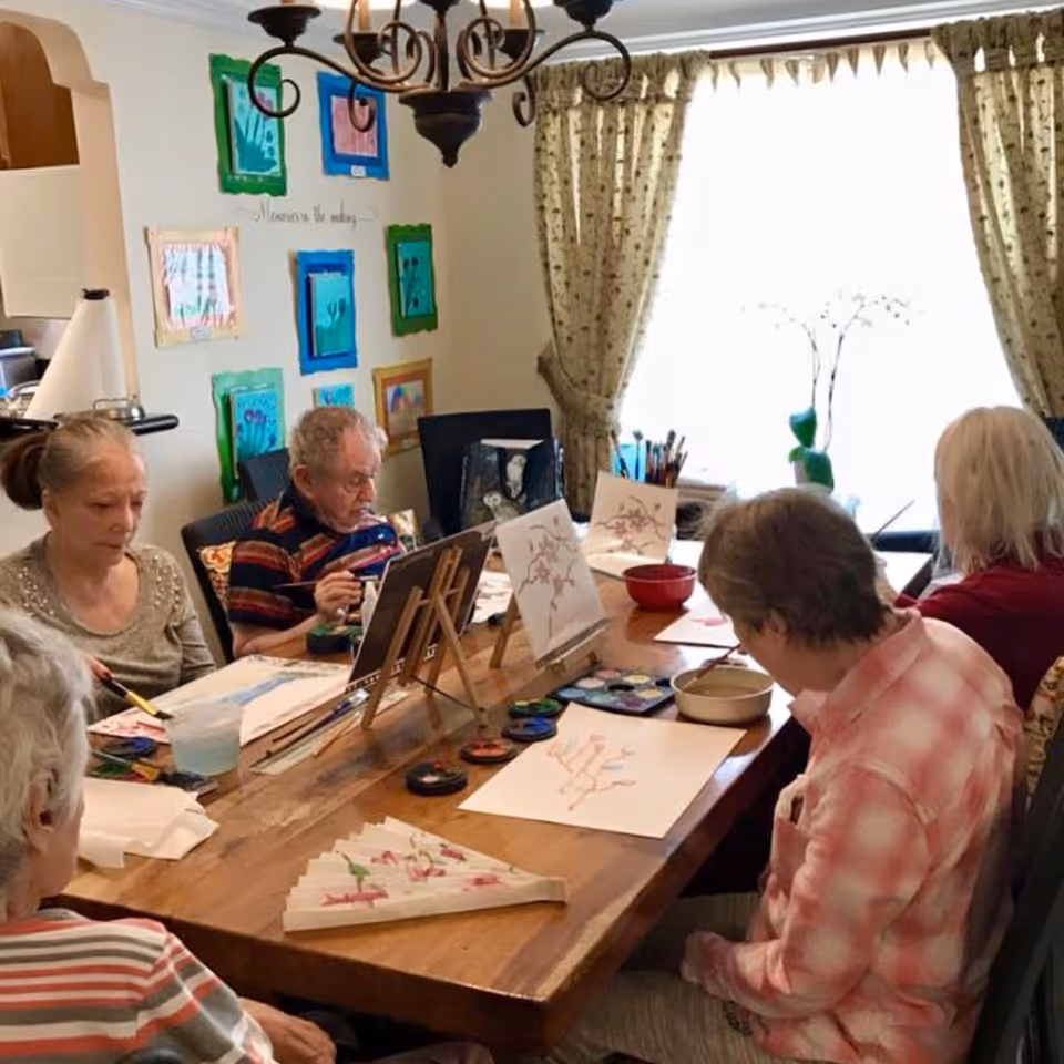 A group of elderly people sitting around a wooden table engaged in painting activities. The table is set with paint palettes, brushes, water containers, and paper with floral designs. The room has a window with patterned curtains letting in natural light, and colorful framed artwork is displayed on the wall.