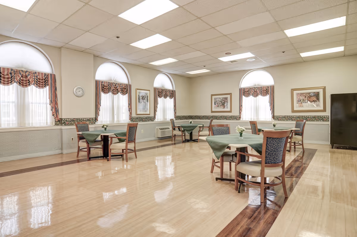 A bright dining room with polished wooden floors and several small round tables covered with green tablecloths. Each table is surrounded by wooden chairs with patterned cushions. The room has large arched windows with sheer white curtains and plaid valances. The walls are decorated with floral wallpaper borders and framed artwork. A clock is mounted on one wall, and there is a black refrigerator in the corner.