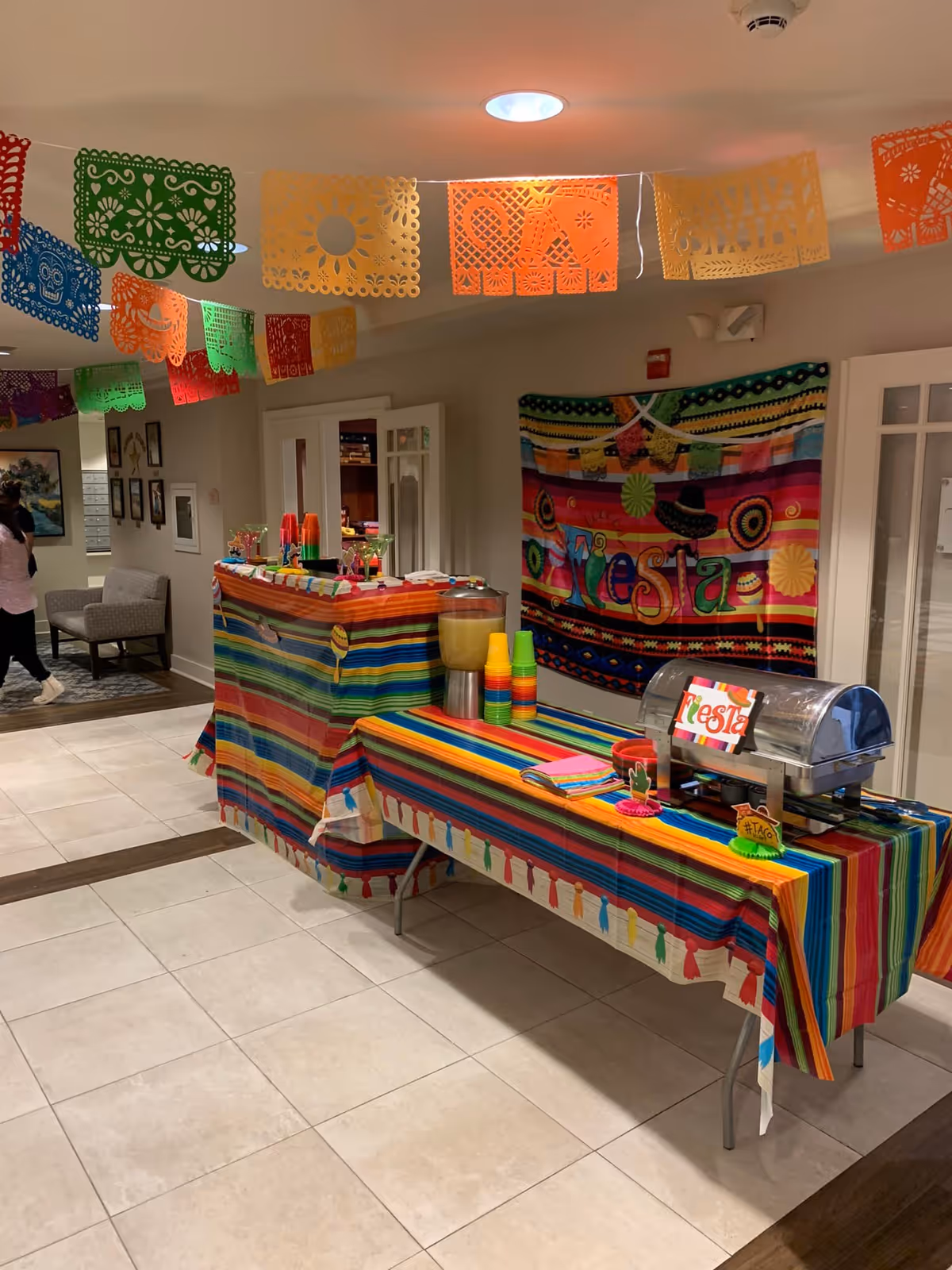 A festive indoor setup with colorful striped tablecloths on two tables arranged in an L shape. One table has a beverage dispenser with stacked colorful plastic cups, and the other has a covered food tray with a sign that says 'Fiesta'. Brightly colored papel picado banners hang from the ceiling, and a vibrant 'Fiesta' banner is displayed on the wall behind the tables. The setting appears to be a common area or hallway in a senior living facility.