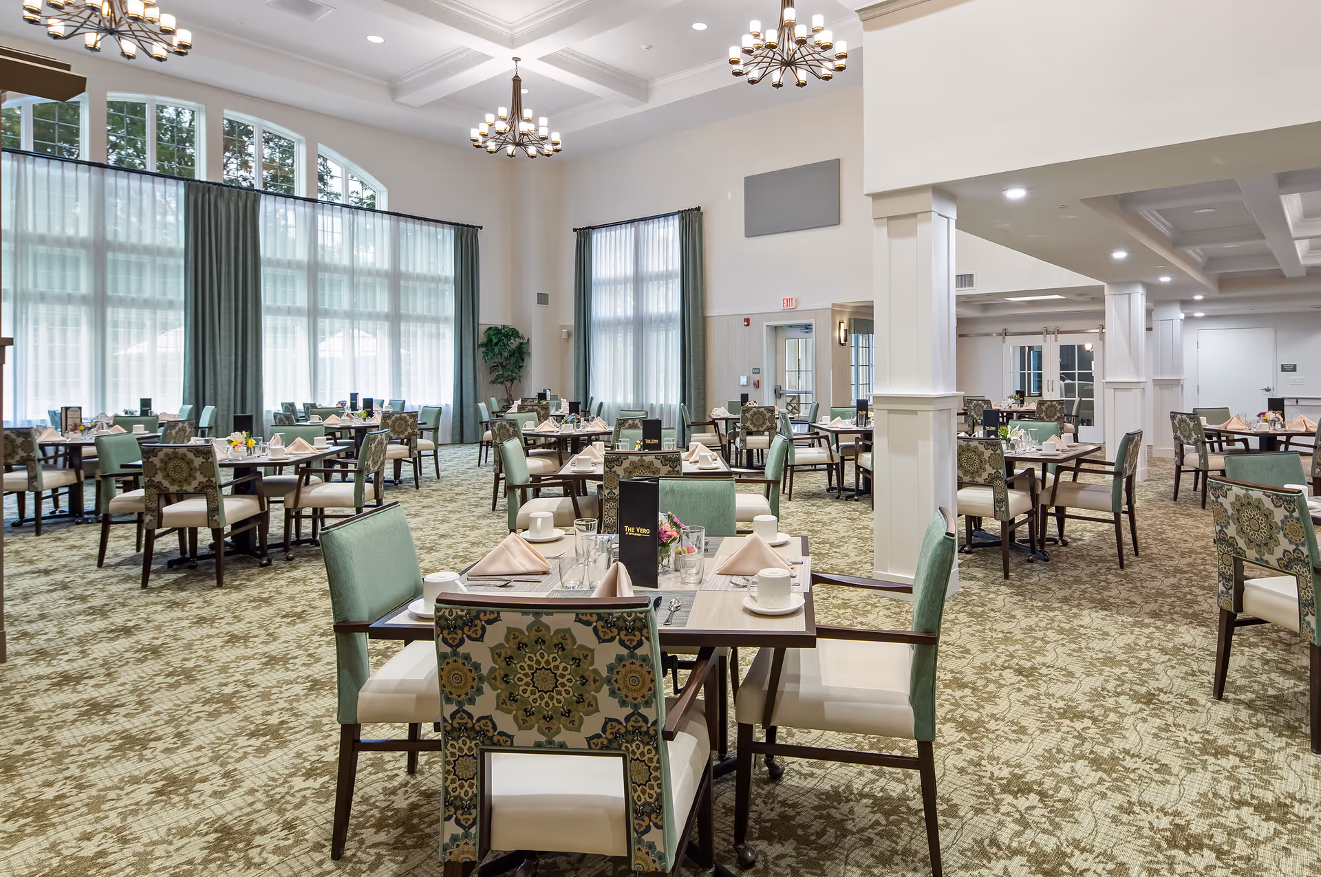 A spacious and well-lit dining room with multiple tables set for meals. Each table has chairs with green and patterned upholstery, beige napkins, cups, glasses, and silverware. Large windows with sheer and green curtains allow natural light to fill the room. The ceiling features elegant chandeliers and a coffered design.
