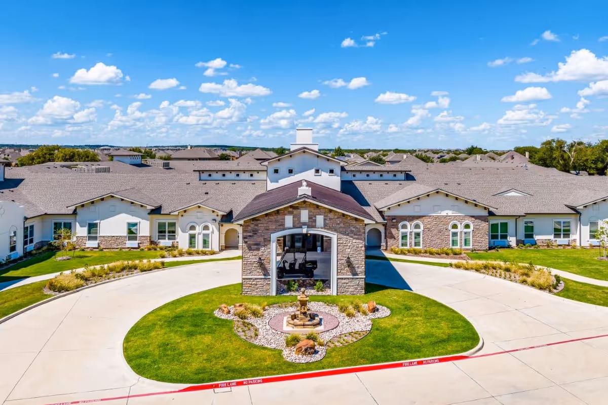 Front exterior view of the Wilshire senior living facility showing a large building with a stone and stucco facade, a circular driveway with a landscaped island featuring a small fountain, and a clear blue sky with scattered clouds.