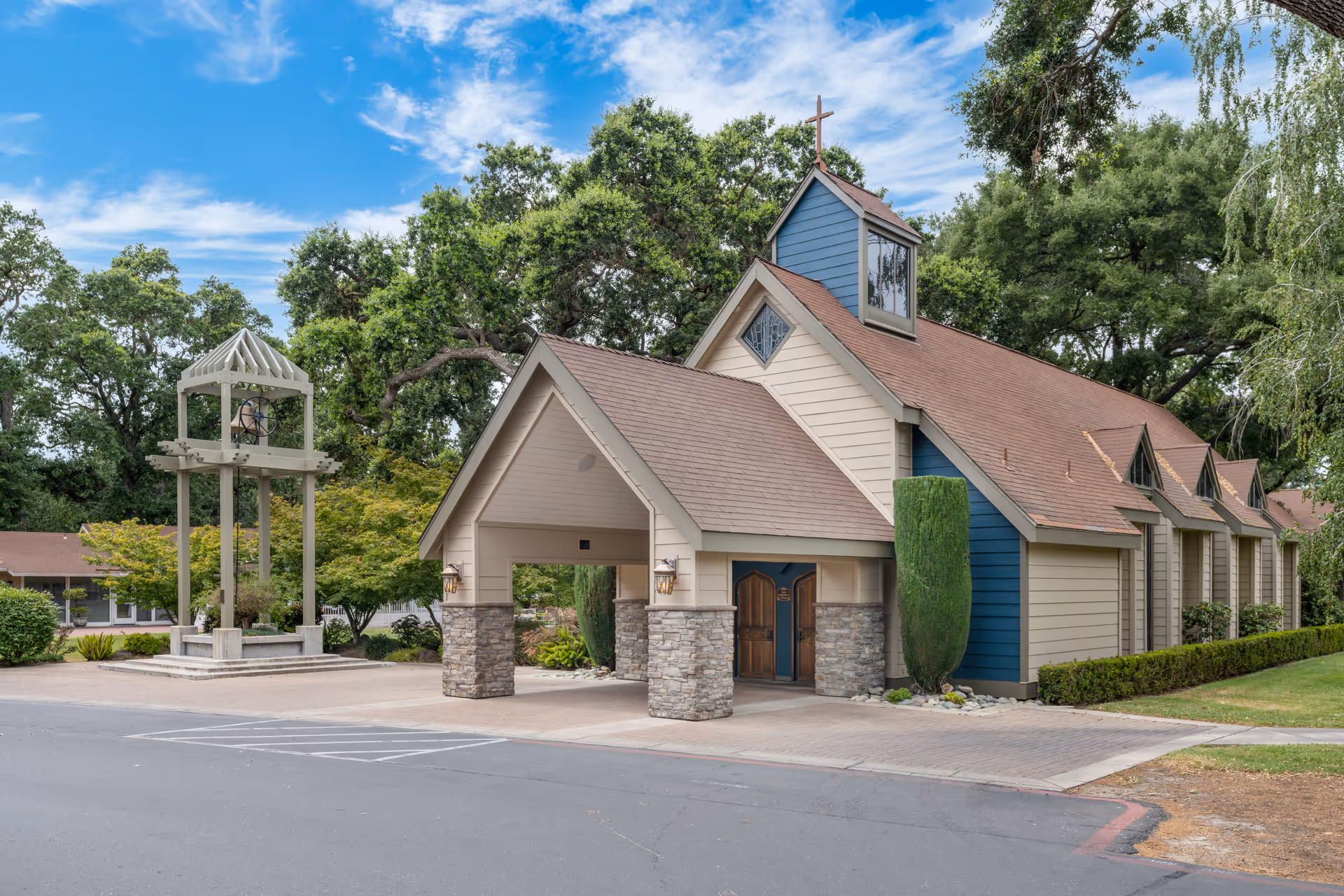 Exterior view of a chapel-like building with a peaked roof and a cross on top, surrounded by trees and greenery. There is a bell tower structure to the left and a paved driveway in front.